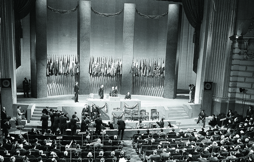 Svart vit bild på FN under bildanden 1945. Bilden visar fullsatta läktare samt några framme vid podiet som fotograferas av pressen.