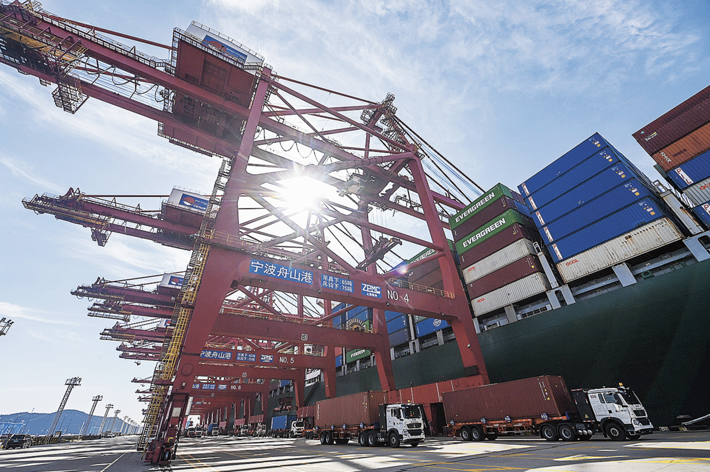 Shanghais hamn där man ser ett stort fartyg och en lastdocka som ska lasta av containrar. Intill står lastbilar uppradade för att ta continrarna vidare. 