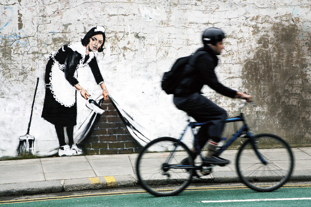 En man cyklar på en gata framför en väggmålning föreställande en kvinna i traditionell städerskeklädsel. Kvinnan drar upp ett skynke som täcker väggen och sopar in skräp bakom det.